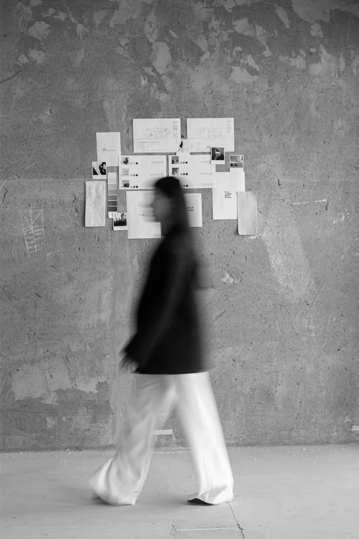 Blurred figure walking past a pinned moodboard of layouts and swatches, concrete studio wall